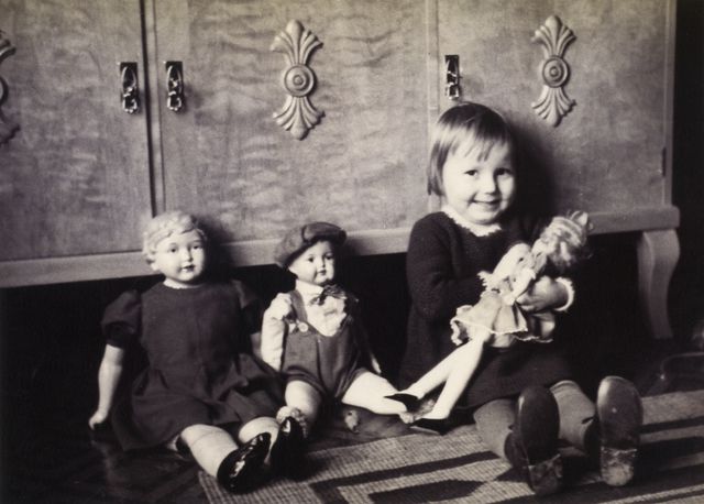 Valokuvassa pieni tyttö pitää sylissään nukkea vierellään kaksi muuta nukkea.