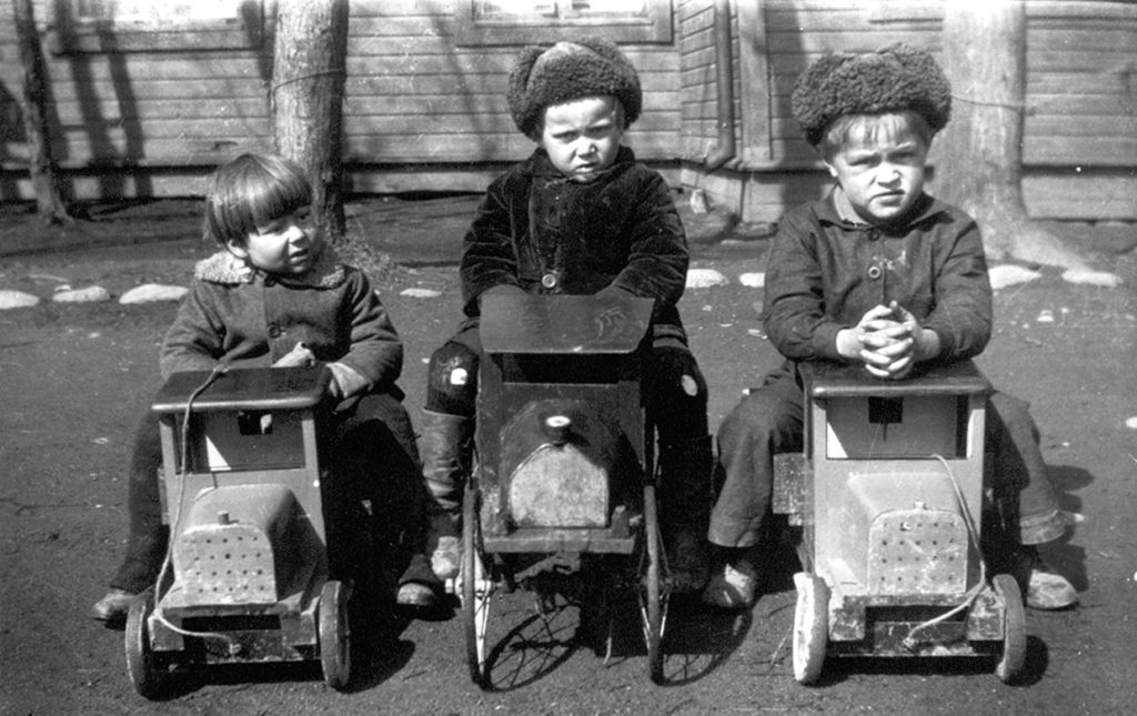 mustavalkoisessa valokuvassa kolme poikaa istuu leluautojen päällä pihalla.