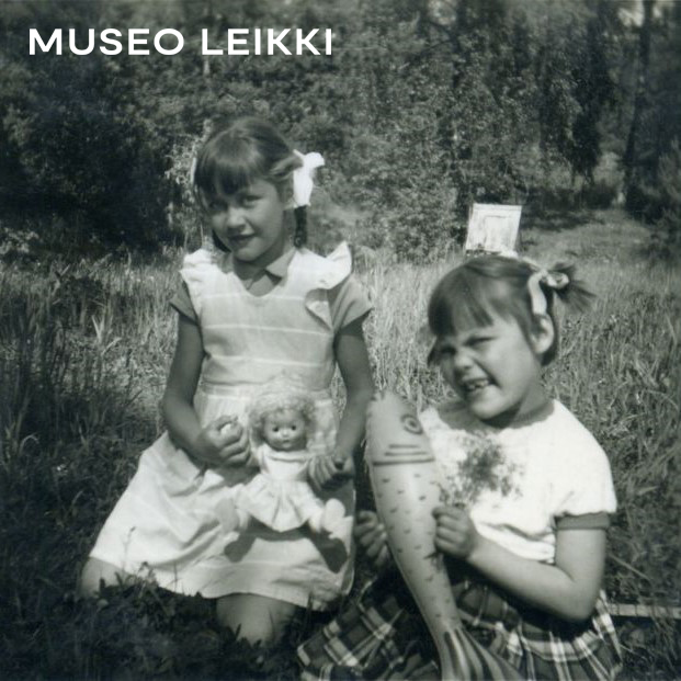 Moilanen Tuula, kuvaaja 1959 Rusettipäiset tytöt istuvat ruohikolla kotipihalla. Kahdeksanvuotias isosisko Pirjo-Liisa pitää sylissään Mirva-nukkea. Kuusivuotiaalla pikkusiskolla Marja-Leenalla on kädessään iso keltainen muovikala ja toisessa kädessä pieni kukkakimppu